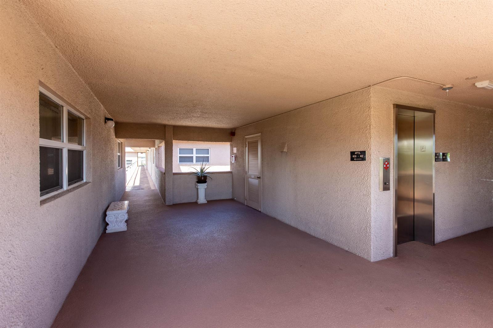 An outdoor apartment hallway features a metal elevator, a small bench, and a potted plant — 2615 NE 1st Court # 411, Boynton Beach FL 33435, BOYNTON BEACH, FL