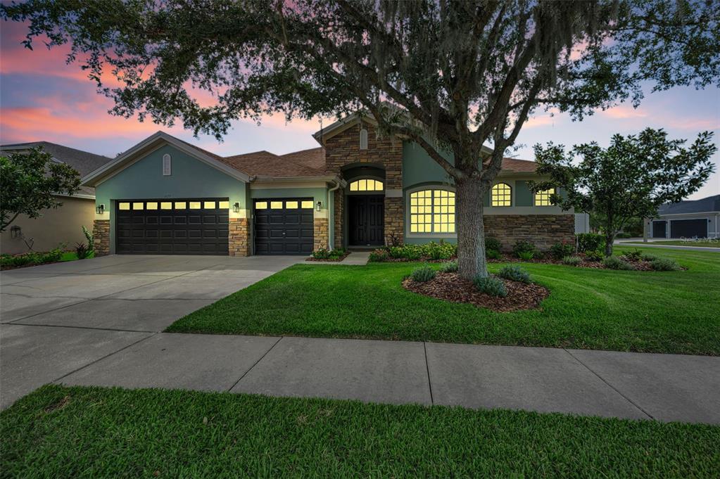 A single-story house with a stone facade, green siding, a three-car garage, and a large tree — 1629 SALMONBERRY STREET, WESLEY CHAPEL, FL