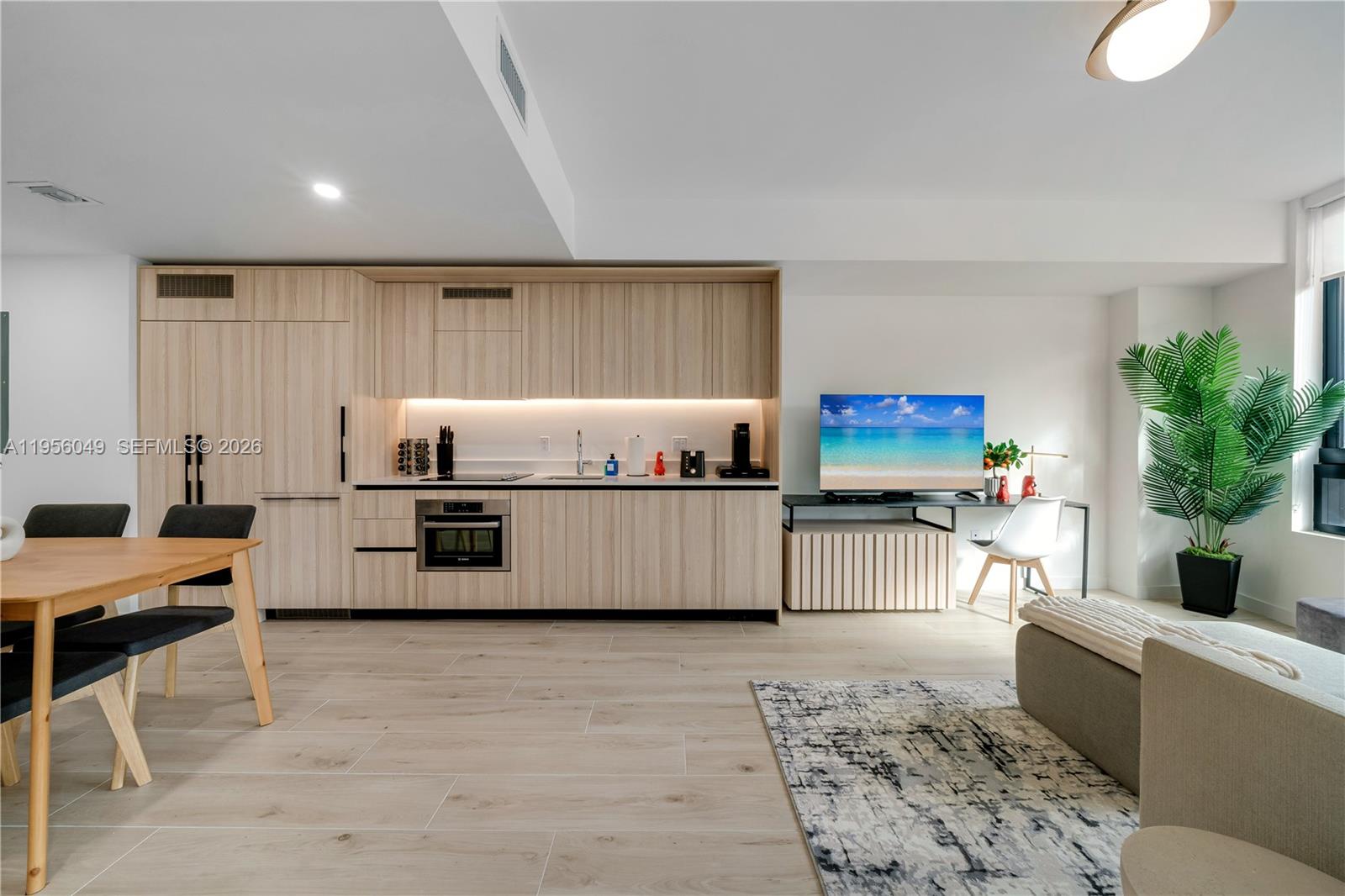A modern kitchen with light wood cabinetry flows into a living area with a television — 225 N Miami Ave # 1707, Miami FL 33128, MIAMI, FL