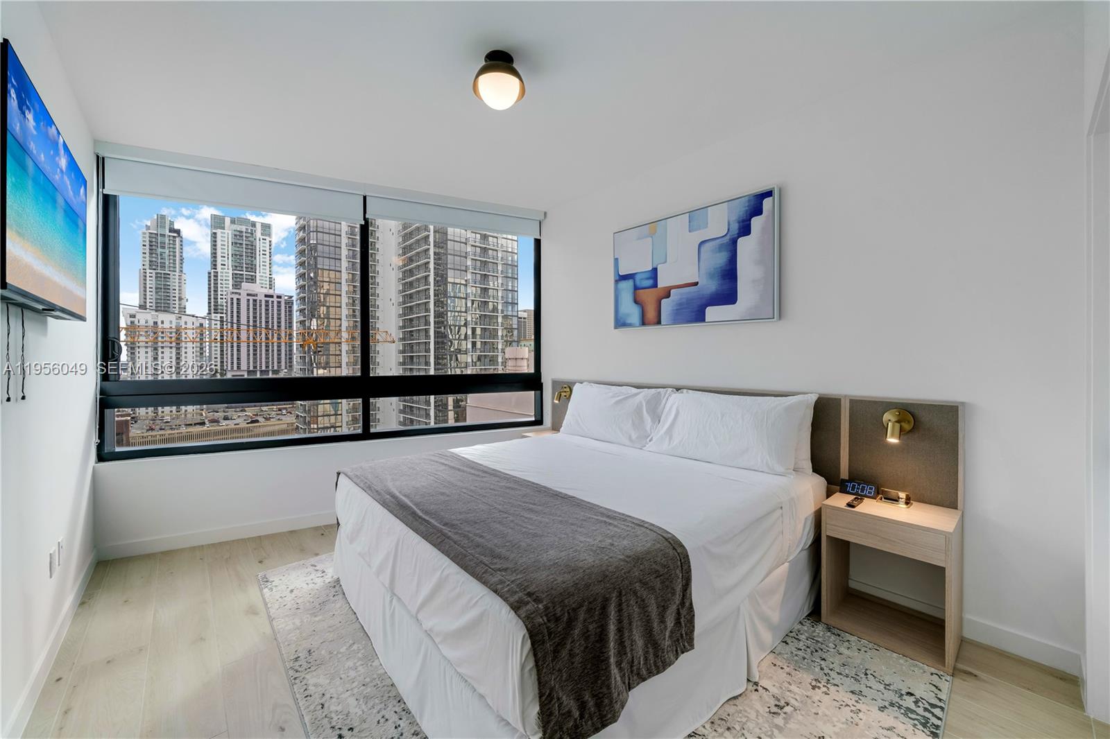 A bedroom features a queen bed, a wall-mounted television, and floor-to-ceiling windows overlooking city skyscrapers — 225 N Miami Ave # 1707, Miami FL 33128, MIAMI, FL