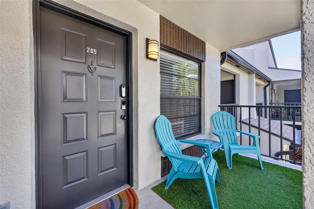 An apartment entrance with a dark door, two turquoise chairs, and artificial turf on a balcony — 2597 COUNTRYSIDE BOULEVARD UNIT 209, CLEARWATER, FL