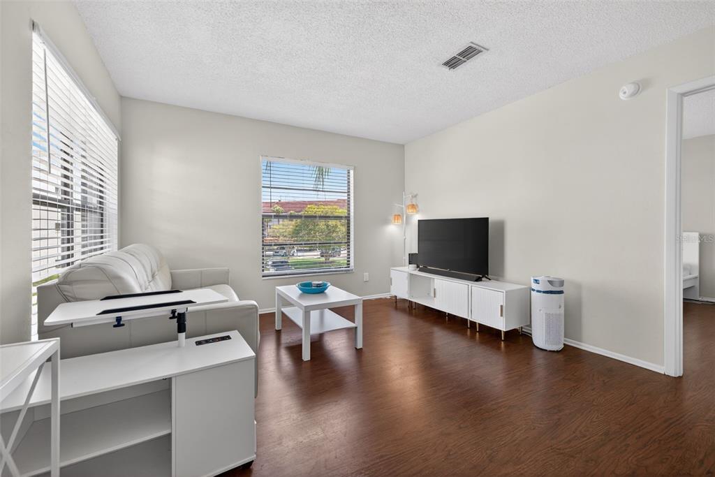 A living room features white furniture, a television, wood-look flooring, and a window with blinds — 2597 COUNTRYSIDE BOULEVARD UNIT 209, CLEARWATER, FL