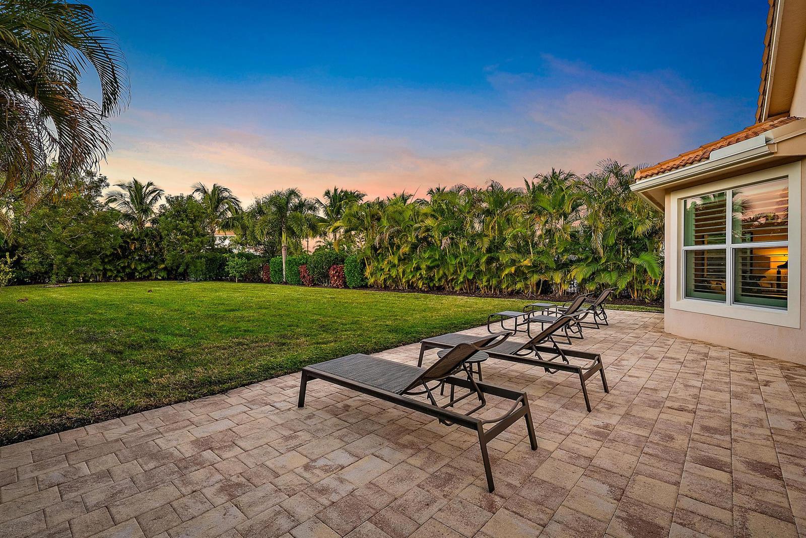 A paved patio features several lounge chairs overlooking a grassy backyard with palm trees — 271 Carina Drive, Jupiter FL 33478, JUPITER, FL