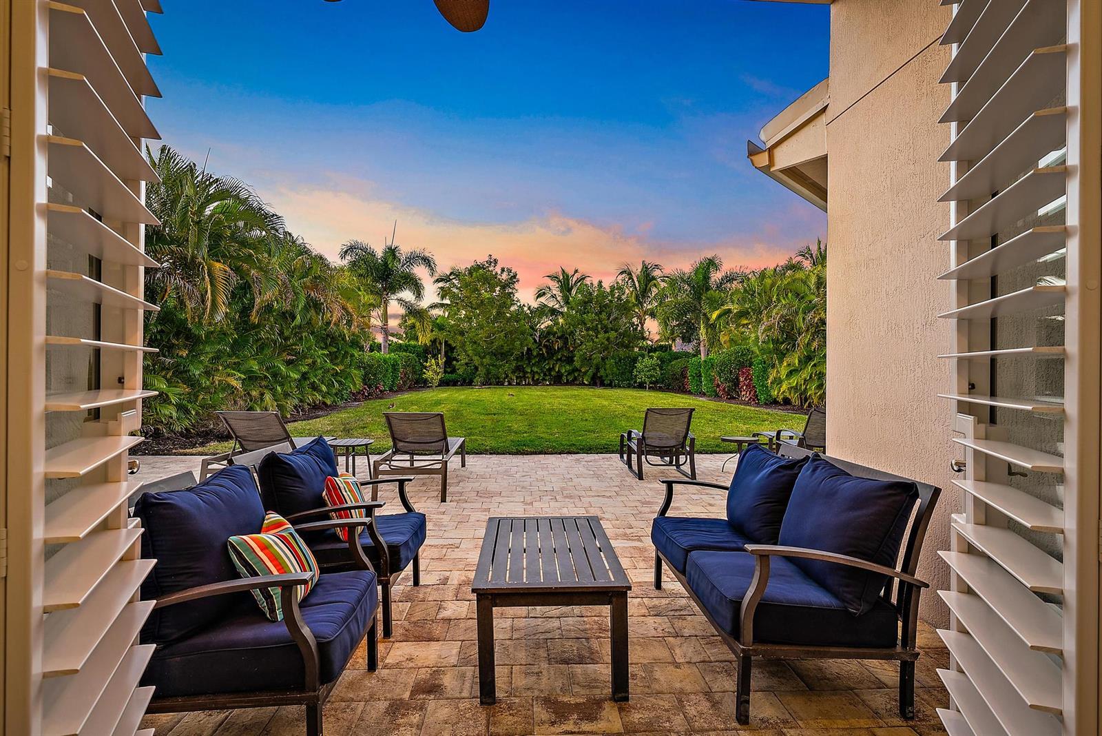 A patio with outdoor furniture overlooks a grassy backyard surrounded by palm trees at sunset — 271 Carina Drive, Jupiter FL 33478, JUPITER, FL