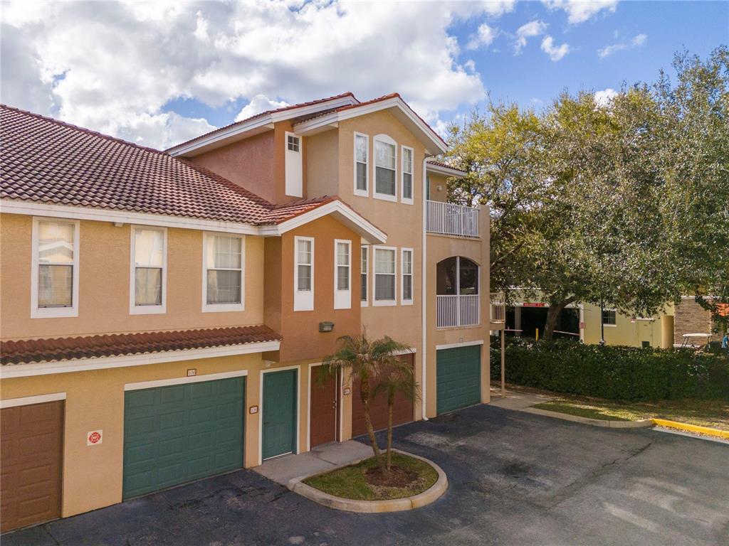 An exterior view of a multi-story tan townhouse with green garage doors and tile roofs — 12102 POPPY FIELD LANE UNIT 107, ORLANDO, FL