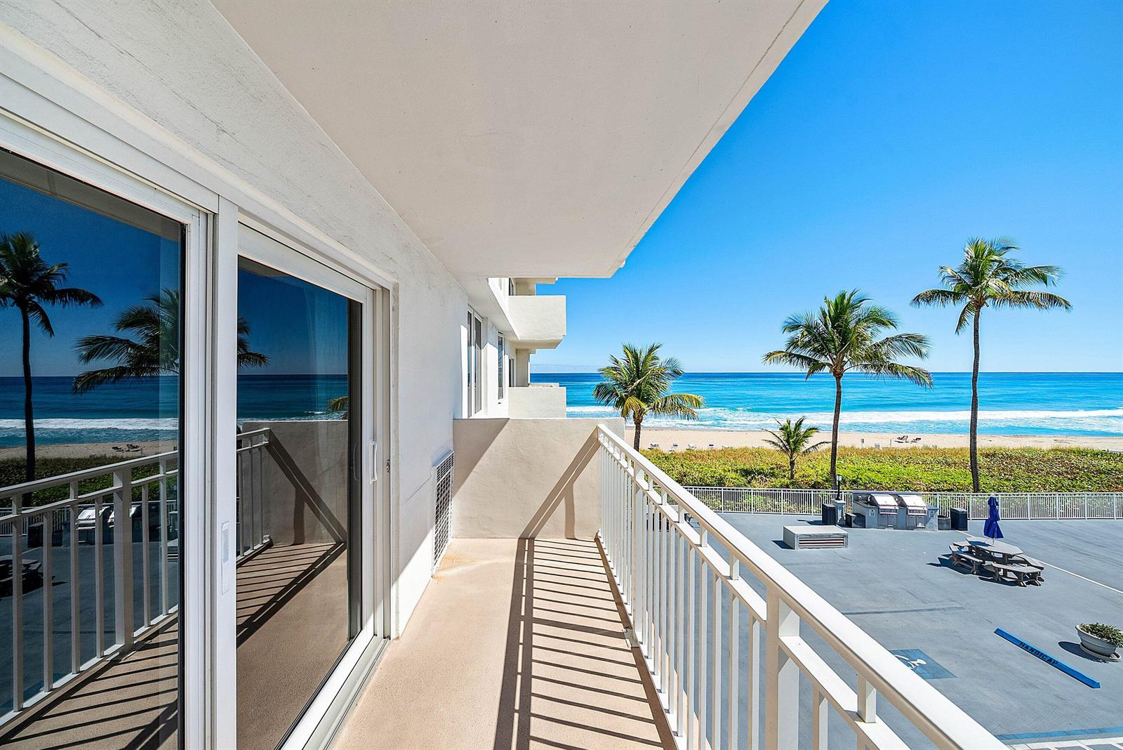 A balcony with a white railing overlooks a beach, palm trees, and a parking area — 3301 S Ocean Boulevard # 207, Highland Beach FL 33487, HIGHLAND BEACH, FL