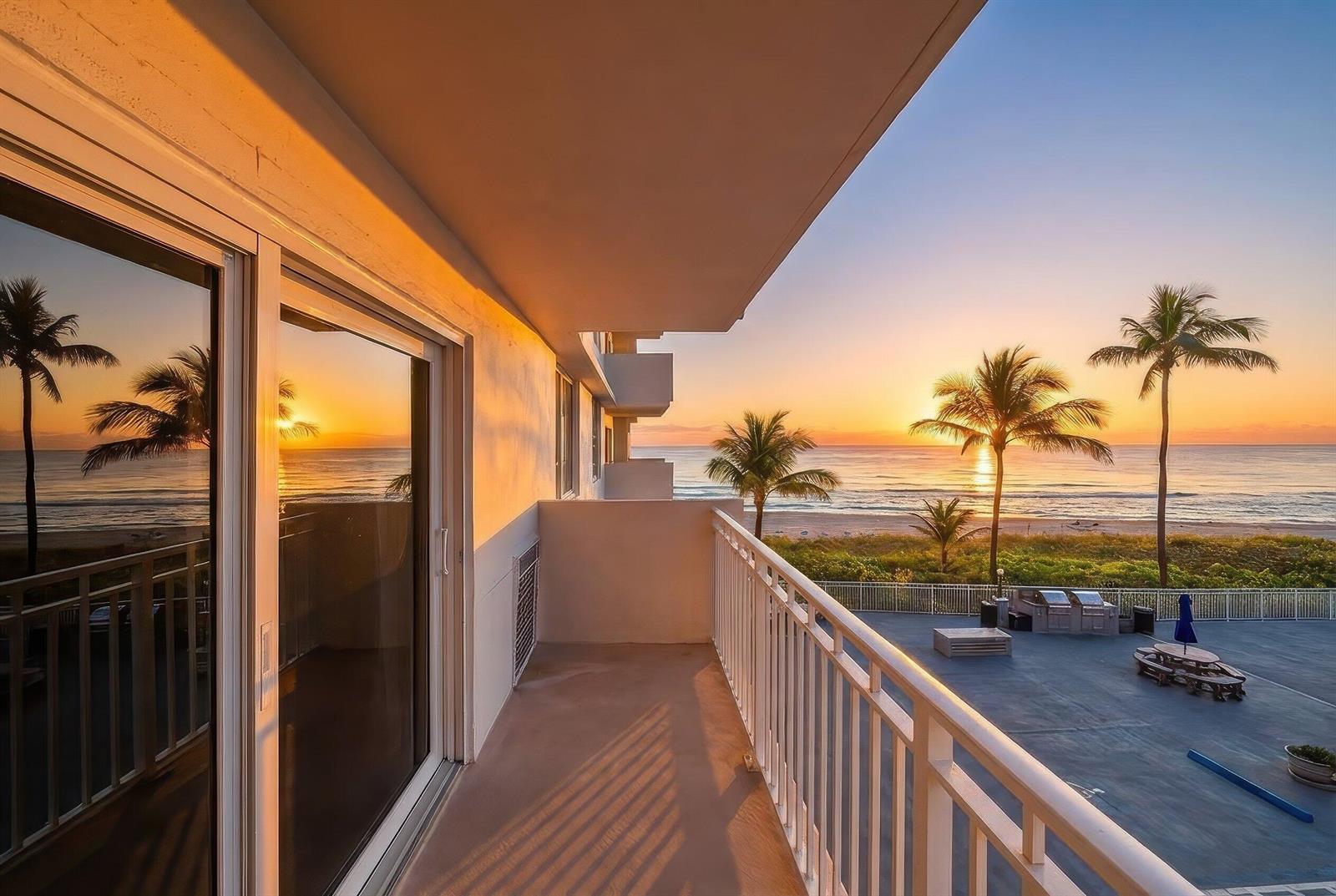 A balcony with a white railing overlooks a beach at sunrise with palm trees — 3301 S Ocean Boulevard # 207, Highland Beach FL 33487, HIGHLAND BEACH, FL