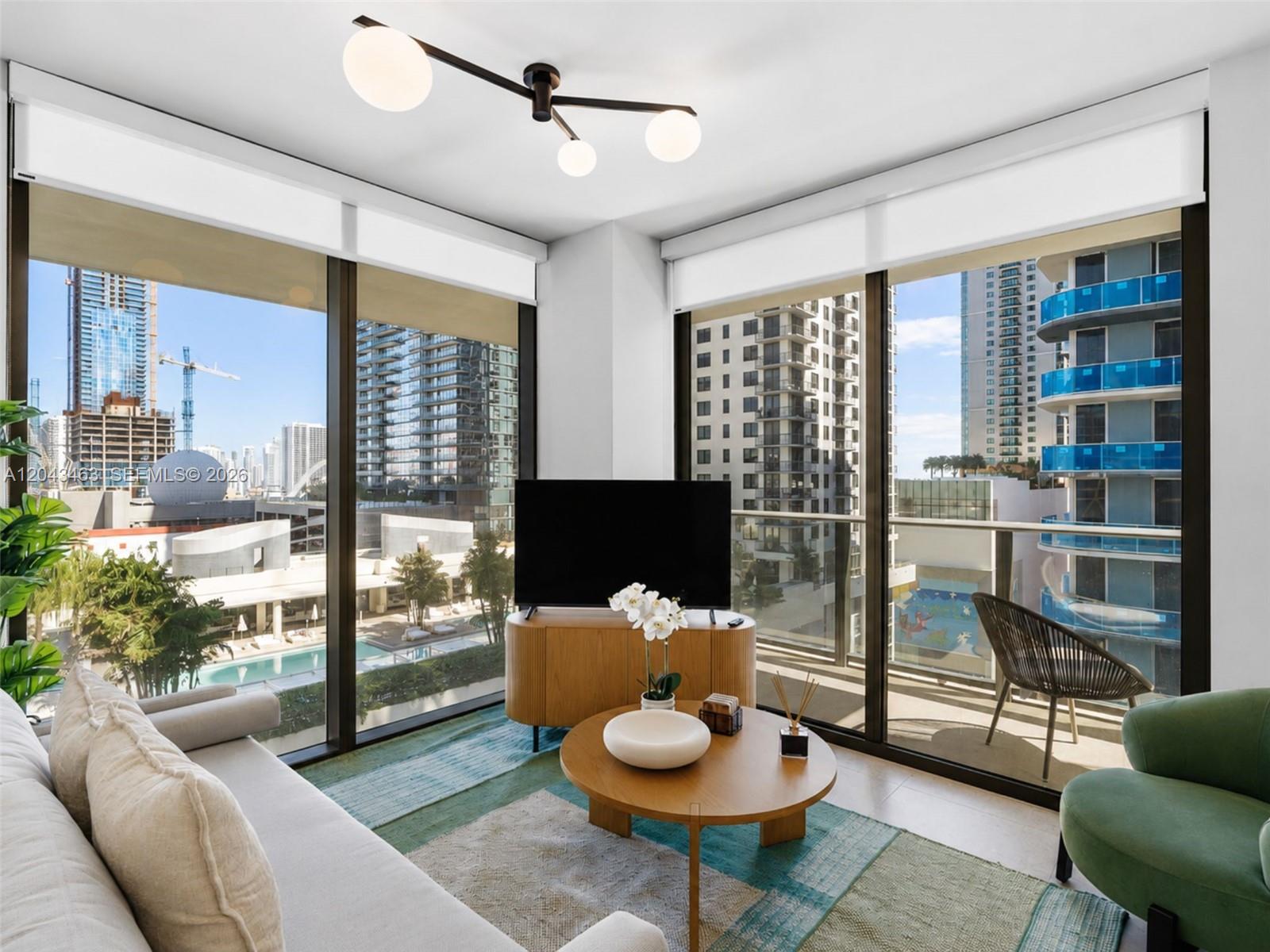 A modern living room features floor-to-ceiling windows, a television on a wooden console, and city views — 11 NE 6th St # 1509, Miami FL 33132, MIAMI, FL