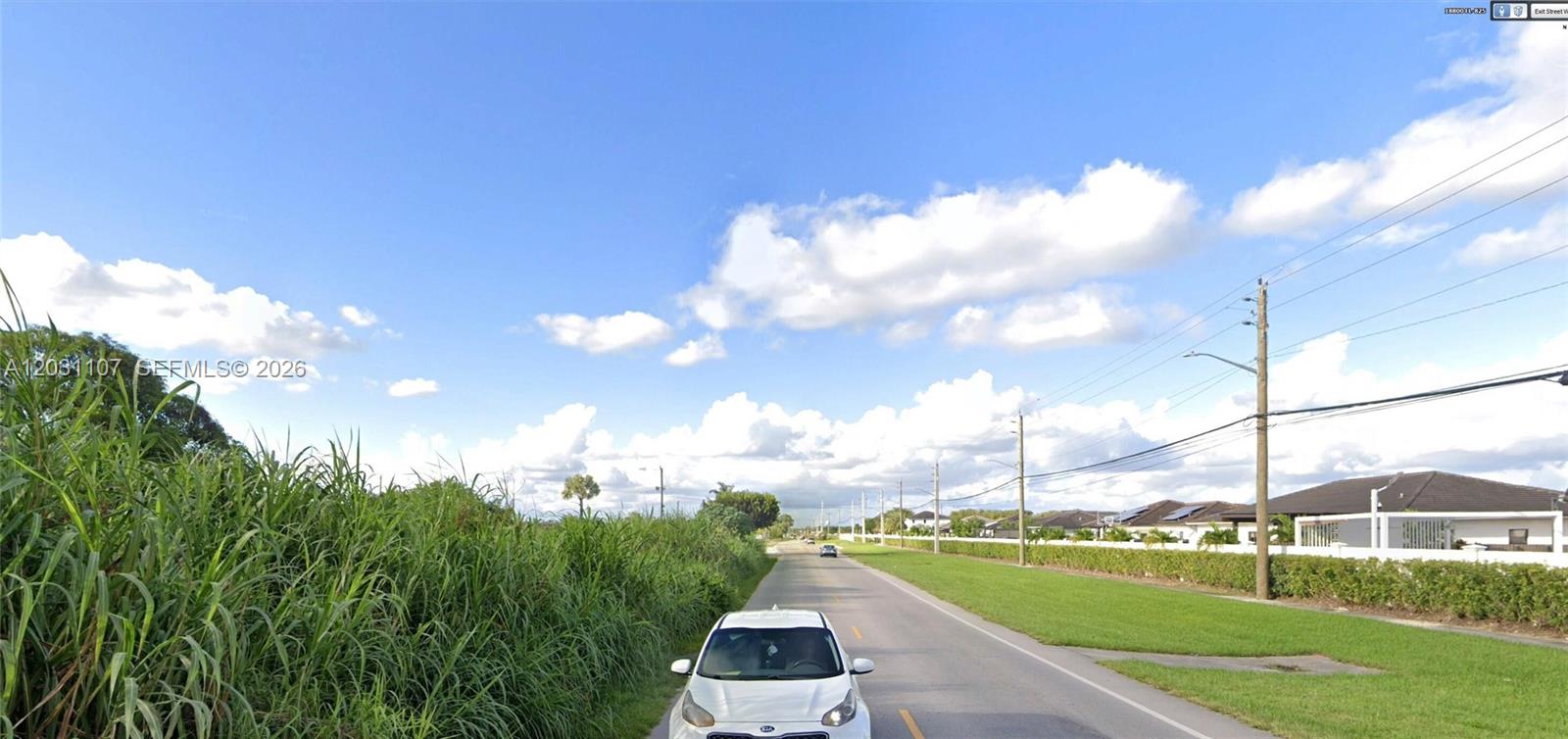 A white car drives down a paved road bordered by tall grass and residential homes — 19000 SW 137th Ave, Miami FL 33177, MIAMI, FL