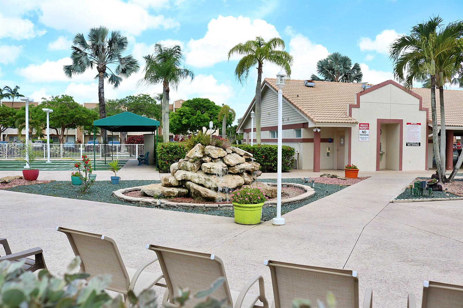 A rock fountain sits in a courtyard with palm trees, lounge chairs, and a building — TAMARAC, FL