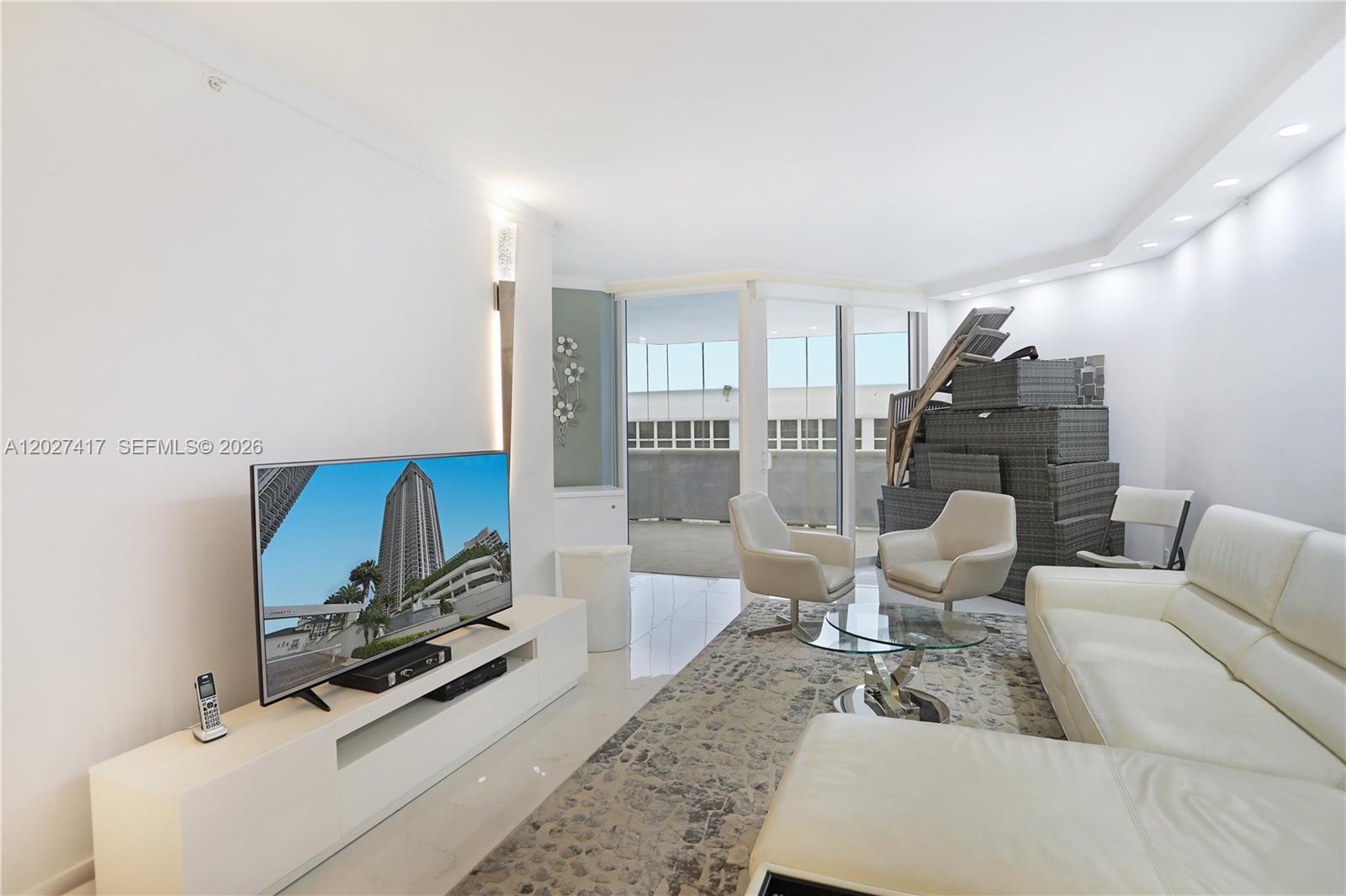 A living room features white leather furniture, a television, and a stack of outdoor chairs — 4775 COLLINS AV # 1804, Miami Beach FL 33140, MIAMI BEACH, FL