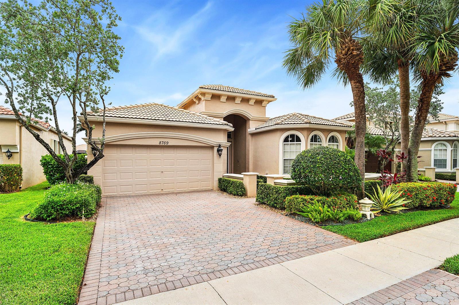 A tan stucco single-story house features a paved driveway, arched windows, and manicured landscaping — 8769 Via Grande E, Wellington FL 33411, WELLINGTON, FL