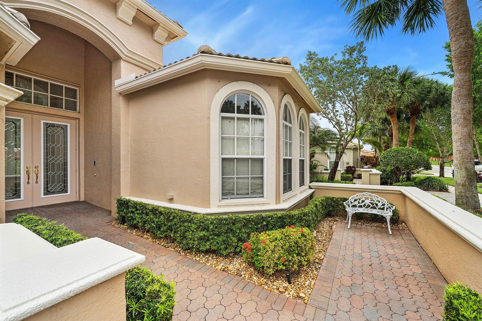 A beige stucco house exterior features a brick walkway, arched windows, and a white bench — 8769 Via Grande E, Wellington FL 33411, WELLINGTON, FL
