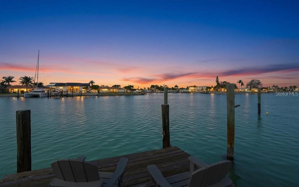 A wooden dock with chairs overlooks a calm bay at sunset with houses in the distance — 510 115TH AVENUE, TREASURE ISLAND, FL