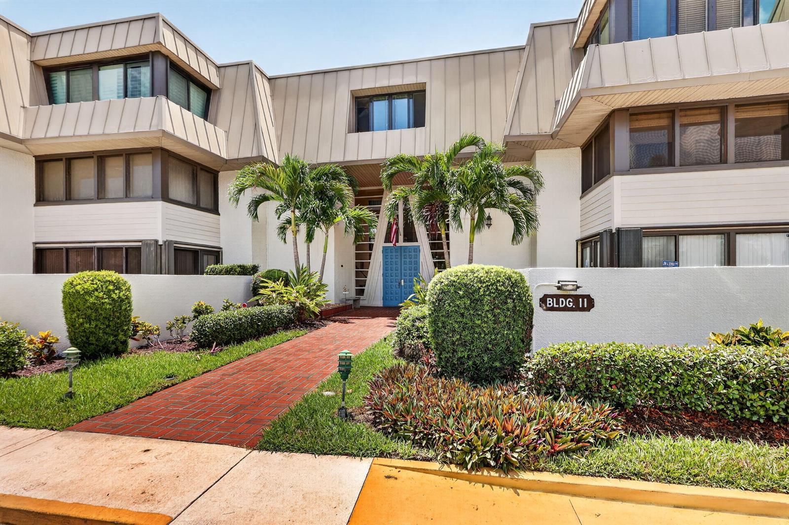 A brick walkway leads to the blue entrance doors of a multi-story building labeled 11 — 1800 SE Saint Lucie Boulevard # 203, Stuart FL 34996, STUART, FL