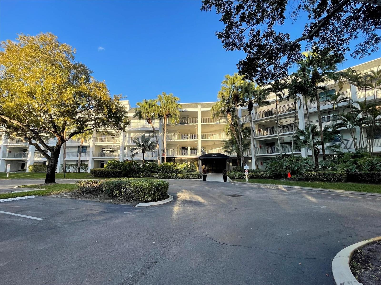 A multi-story white apartment building with balconies and palm trees stands behind a paved driveway — 16141 Blatt Boulevard # 408, Weston FL 33326, WESTON, FL