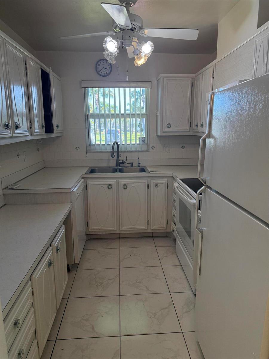 A galley-style kitchen features white cabinets, light countertops, tile flooring, and a window above the sink — 211 Country Lane # A, Boynton Beach FL 33435, BOYNTON BEACH, FL