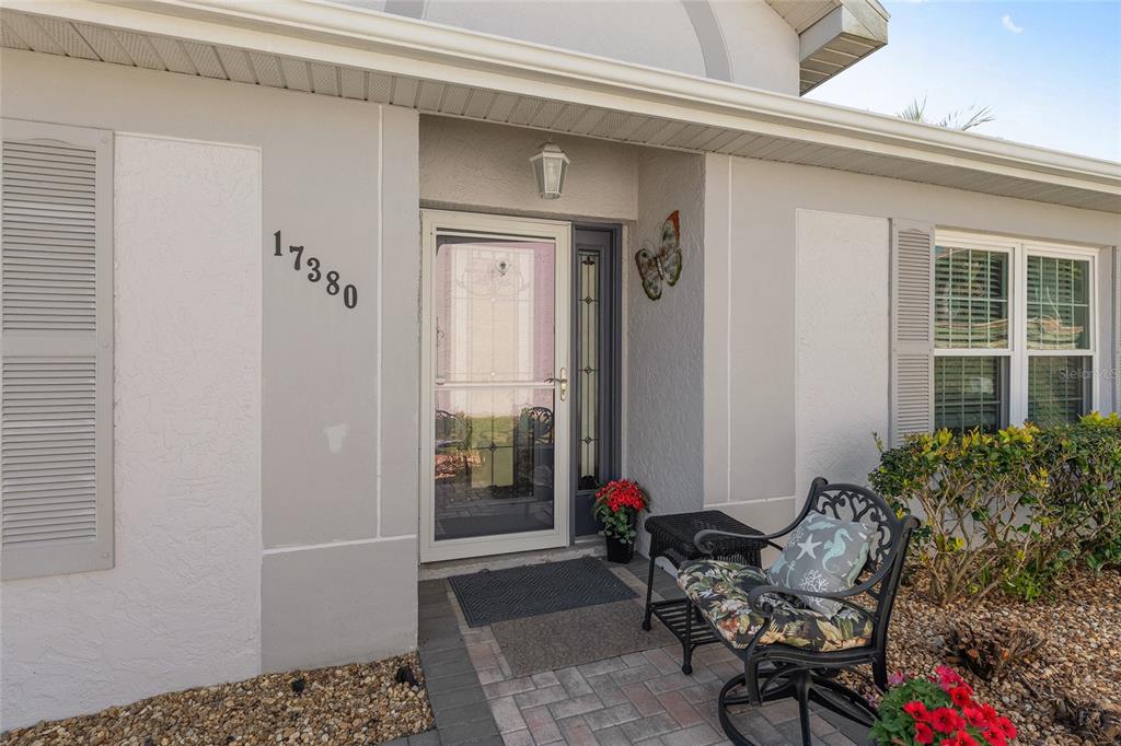 The exterior entrance of a house features a storm door, address numbers, and a chair — 17380 SE 111TH COURT, SUMMERFIELD, FL
