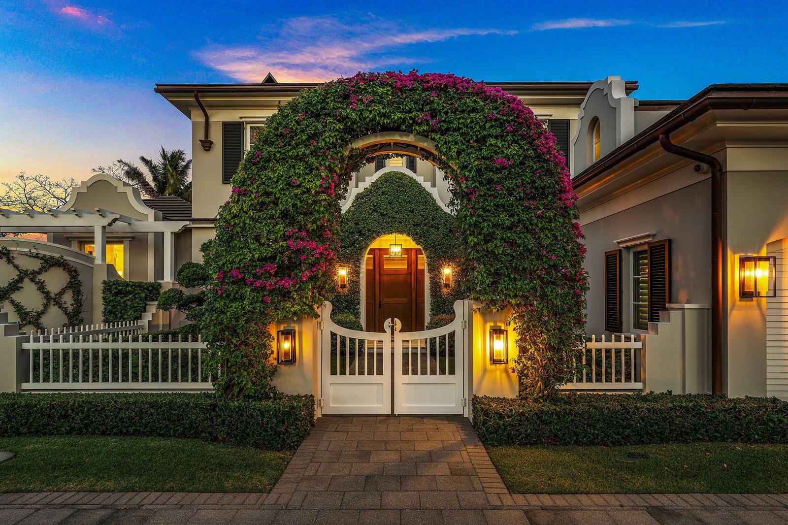 A white gate sits beneath a large flowering archway leading to a house entrance at dusk — 2530 Avenue Au Soleil, Gulf Stream FL 33483, GULF STREAM, FL