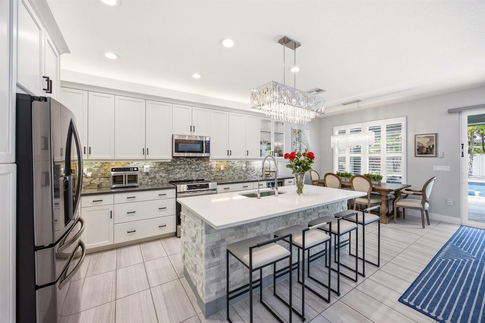 A modern kitchen features white cabinetry, a stone-faced island with bar seating, and tile flooring — 15388 Destiny Drive, Delray Beach FL 33446, DELRAY BEACH, FL