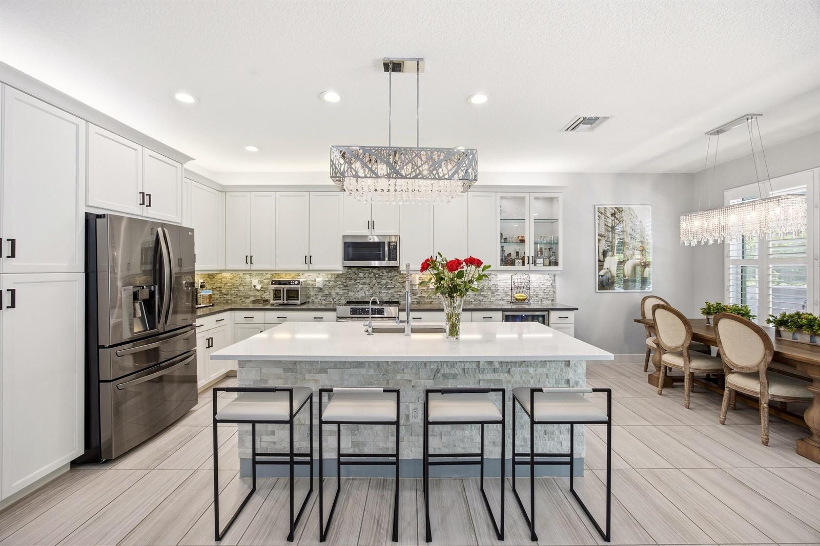 A modern kitchen features white cabinetry, a stone-faced island with bar seating, and crystal chandeliers — 15388 Destiny Drive, Delray Beach FL 33446, DELRAY BEACH, FL