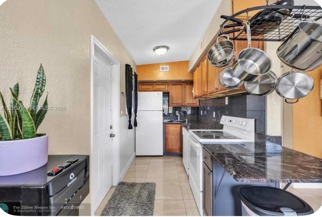 A galley kitchen features wood cabinets, dark granite countertops, white appliances, and a hanging pot rack — 4720 NE 14th Ave, Pompano Beach FL 33064, POMPANO BEACH, FL