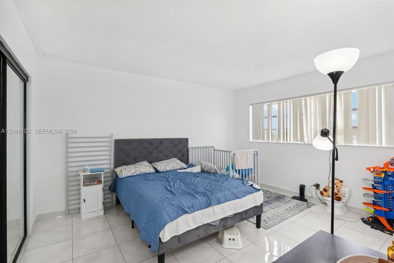 A bedroom features a gray tufted bed, tile flooring, a floor lamp, and a window — HALLANDALE BEACH, FL