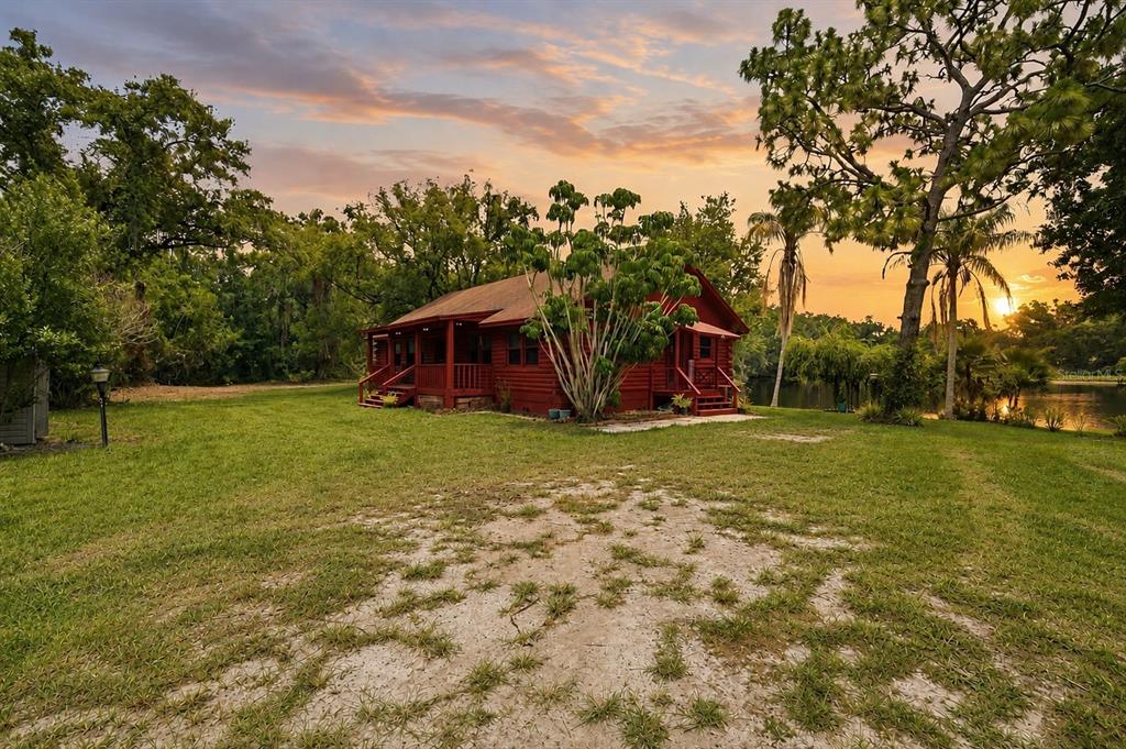 A red log cabin sits on a grassy lawn near a lake at sunset — 6409 CHISWICK COURT, NEW PORT RICHEY, FL