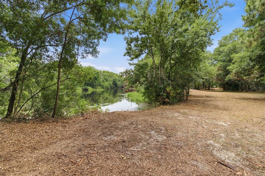 A dirt clearing overlooks a calm lake surrounded by dense trees and foliage — 6409 CHISWICK COURT, NEW PORT RICHEY, FL