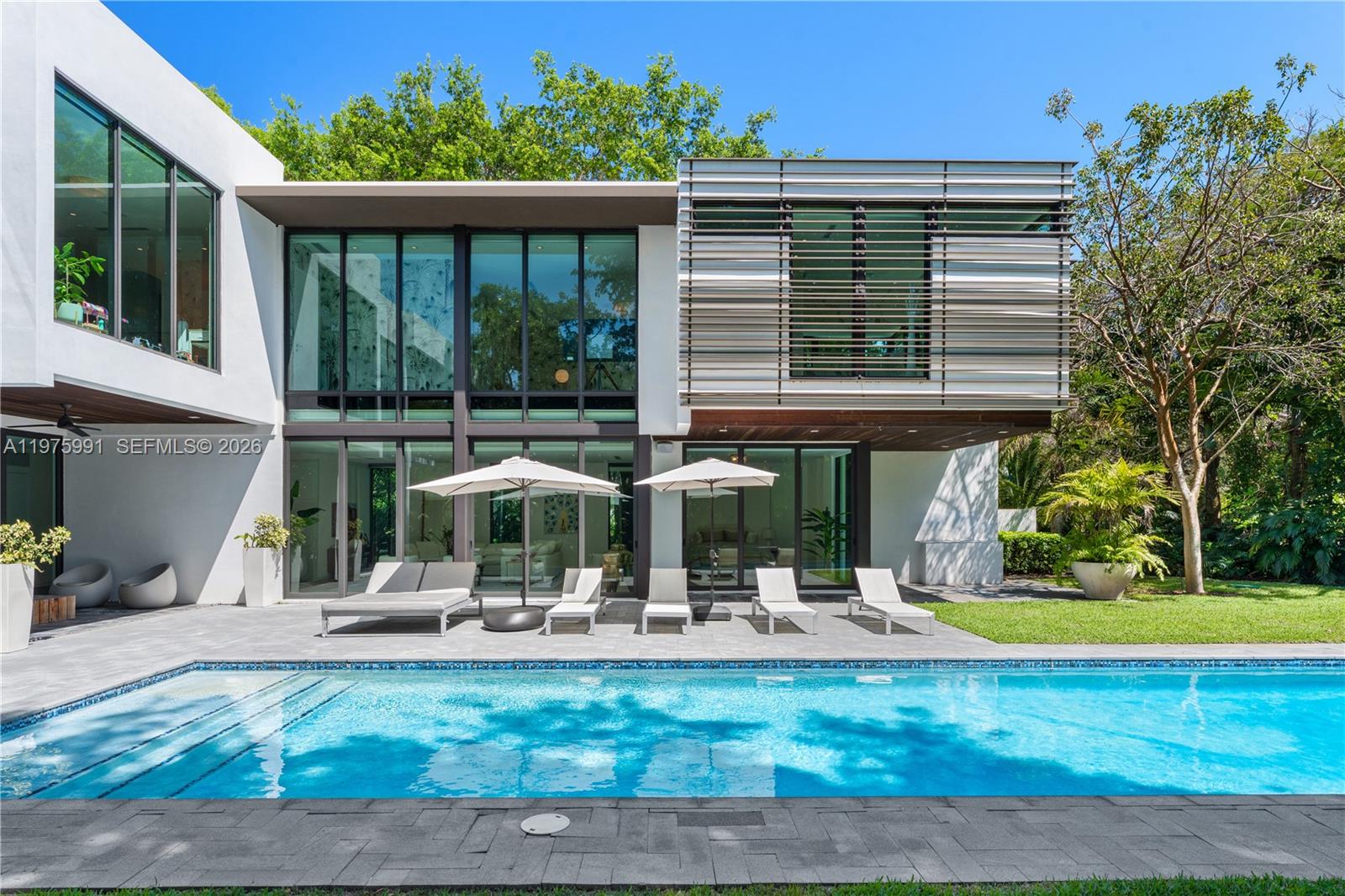 A modern two-story house with a swimming pool, lounge chairs, and large glass windows — 12220 Rock Garden Ln, Pinecrest FL 33156, PINECREST, FL