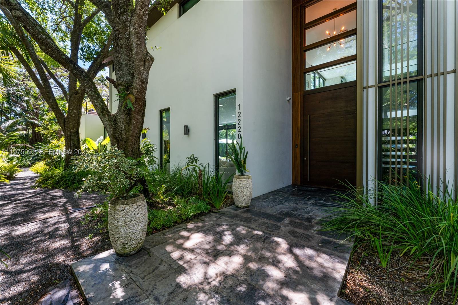 The entrance of a modern home features a large wooden door, stone flooring, and landscaping — 12220 Rock Garden Ln, Pinecrest FL 33156, PINECREST, FL