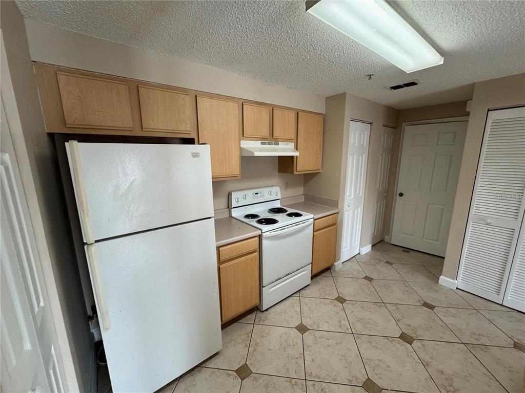 A kitchen features light wood cabinets, white appliances, and beige tile flooring — 10831 WINDSOR WALK DRIVE UNIT 1210, ORLANDO, FL