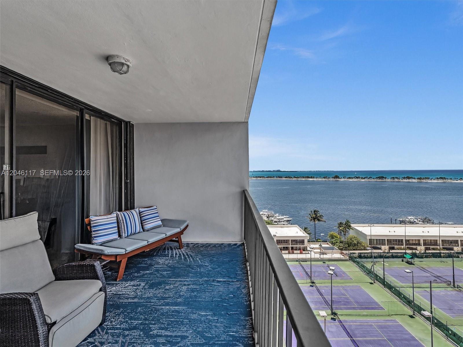 A balcony with outdoor furniture overlooks tennis courts and a bay under a blue sky — 1901 Brickell Ave # B1203, Miami FL 33129, MIAMI, FL