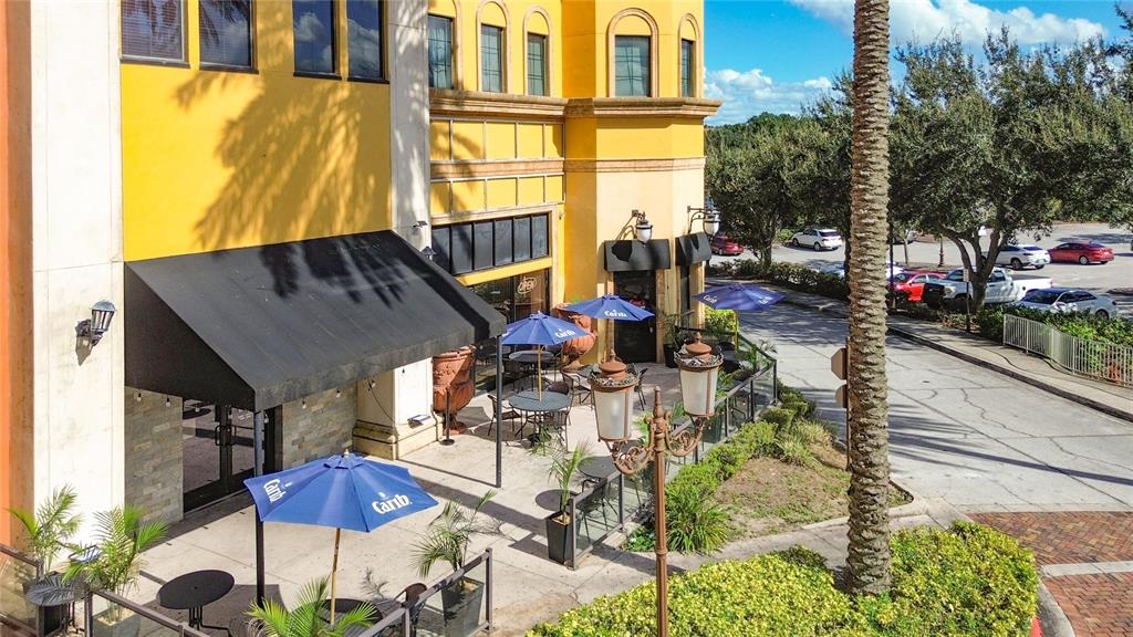 An outdoor patio with tables and blue umbrellas sits beside a yellow commercial building — 6996 PIAZZA GRANDE AVENUE UNIT 100, ORLANDO, FL