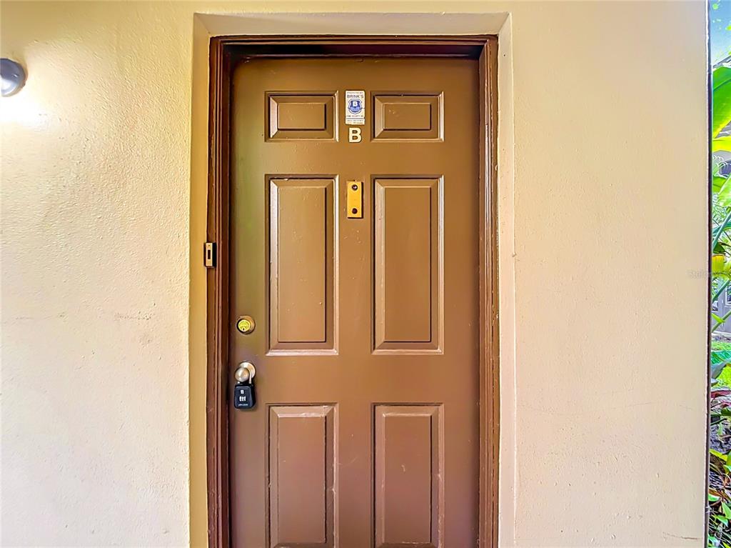 A brown front door with a security system sticker and a lockbox is set in a wall — 11805 RAINTREE LAKE LN UNIT B, TEMPLE TERRACE, FL