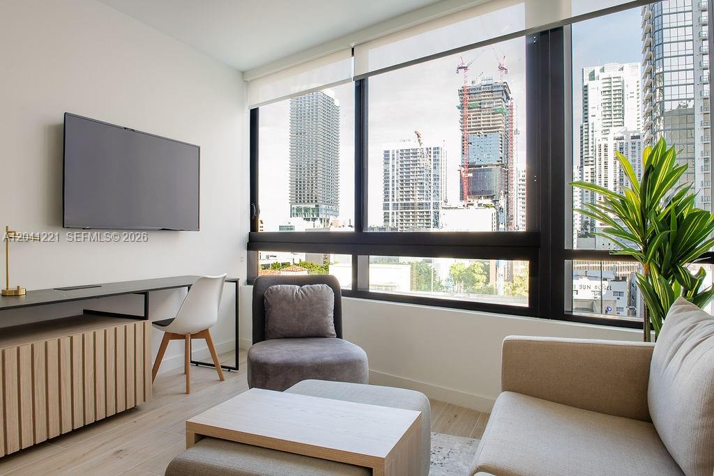 A modern living room features a desk, television, seating, and large windows overlooking city construction — 225 N Miami Ave # 1804, Miami FL 33128, MIAMI, FL