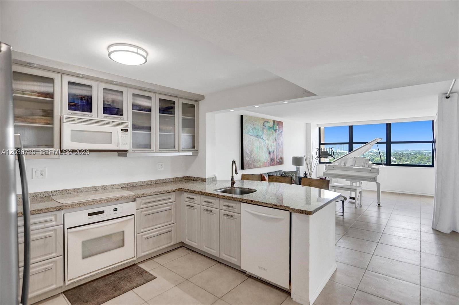 A kitchen with light-colored cabinets and granite countertops opens into a living area with a piano — 2555 Collins Ave # 1508, Miami Beach FL 33140, MIAMI BEACH, FL