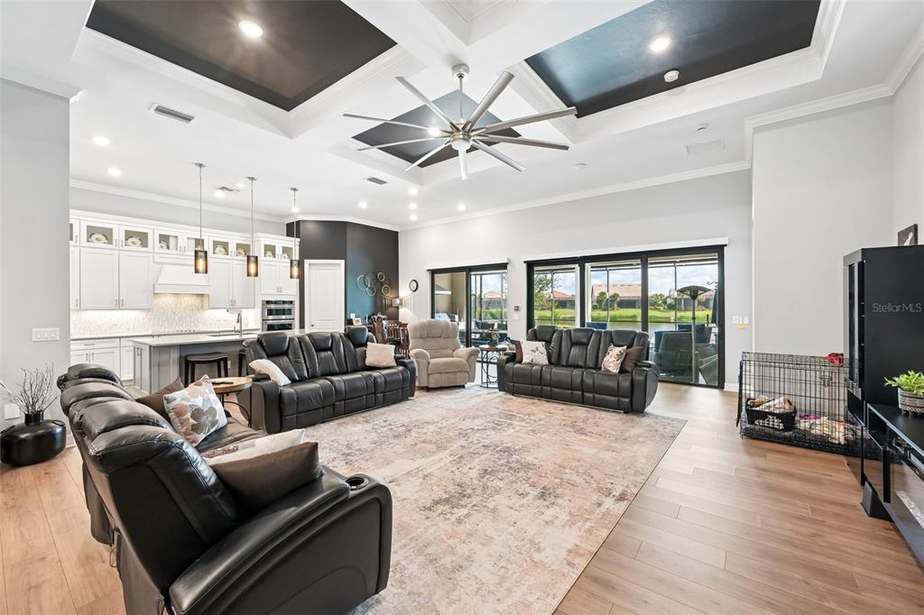A spacious open-concept living room features a coffered ceiling, black leather furniture, and sliding glass doors — 13356 VERANDI STREET, VENICE, FL