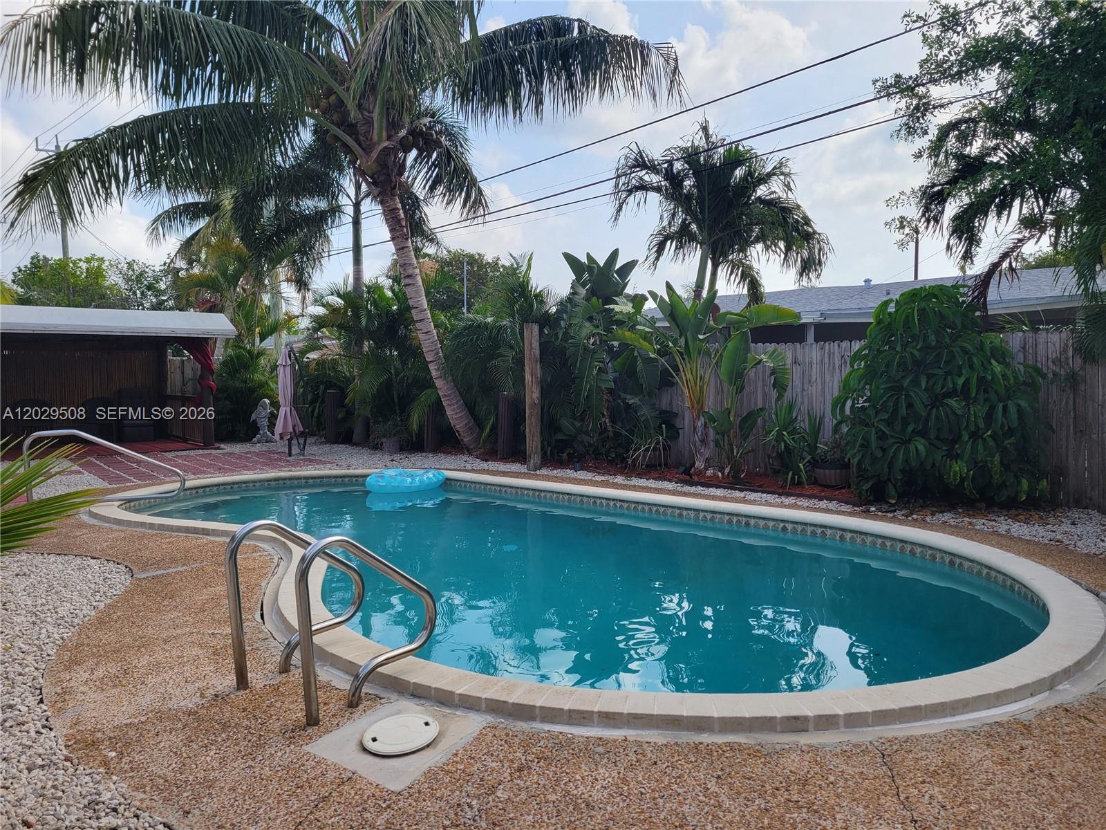 A backyard features an oval swimming pool surrounded by a pebble deck and tropical landscaping — 2560 NE 8th Ter, Pompano Beach FL 33064, POMPANO BEACH, FL