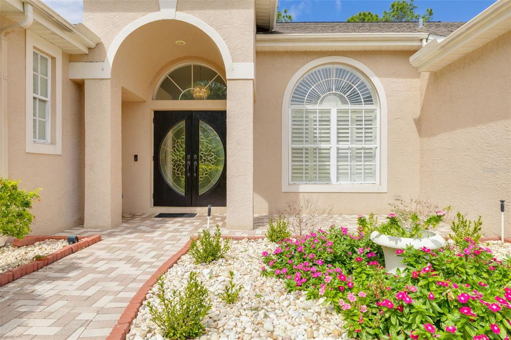 A beige stucco house exterior features a double front door, arched window, and landscaped garden — 3713 SIENA LANE, PALM HARBOR, FL