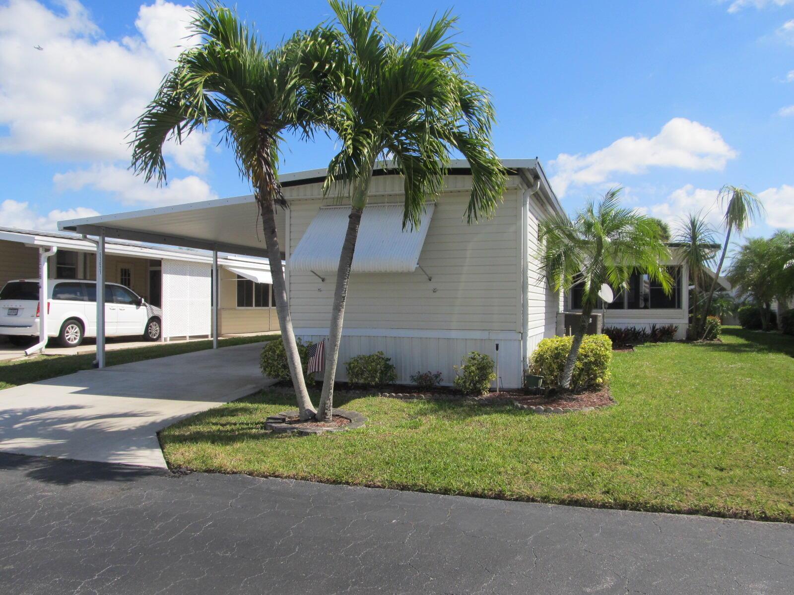 A single-story mobile home with a carport, palm trees, and a lawn under a blue sky — 53021 Del Rio Bay, Boynton Beach FL 33436, BOYNTON BEACH, FL