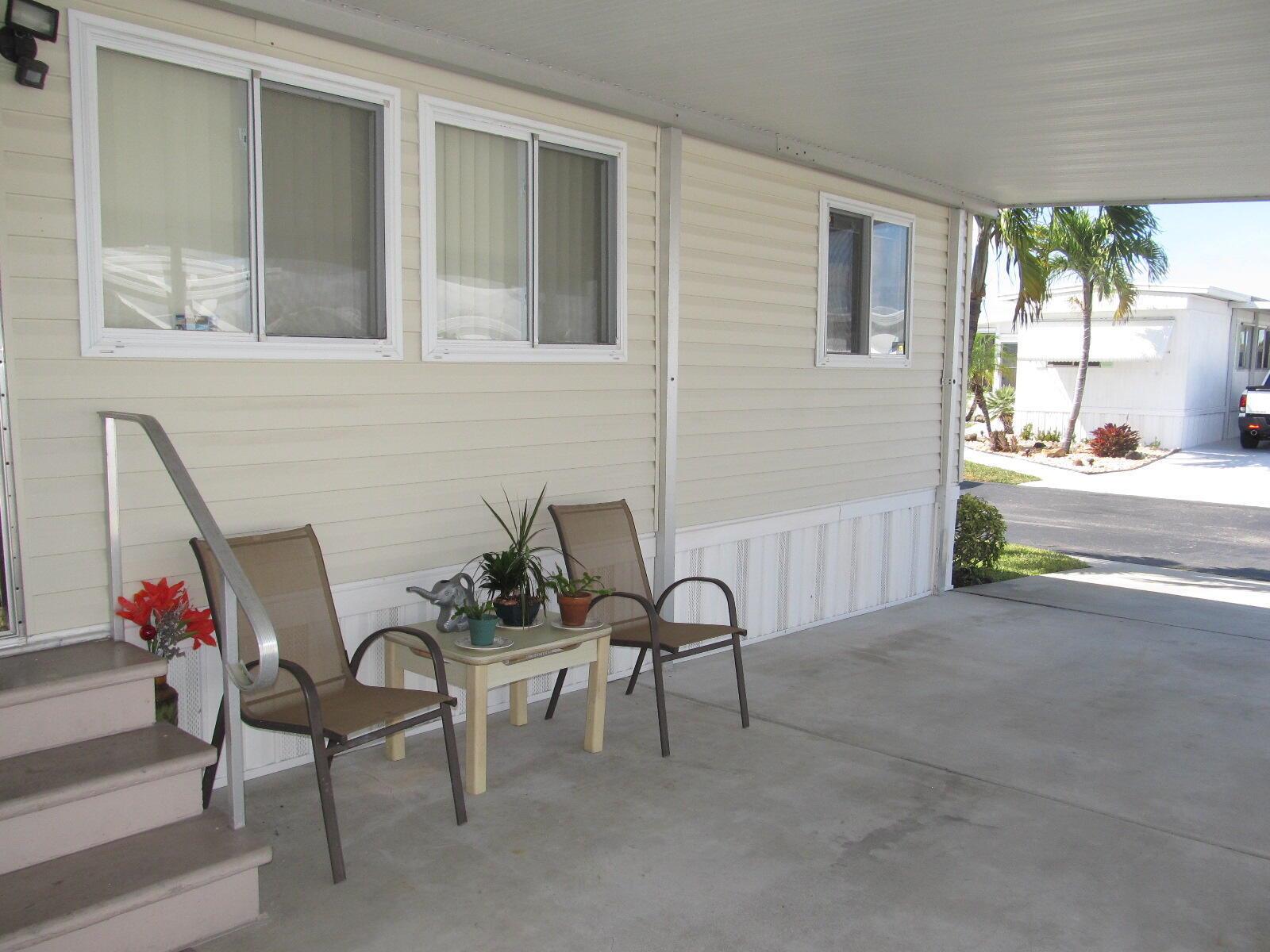 A beige mobile home exterior features a covered concrete patio with two chairs and plants — 53021 Del Rio Bay, Boynton Beach FL 33436, BOYNTON BEACH, FL
