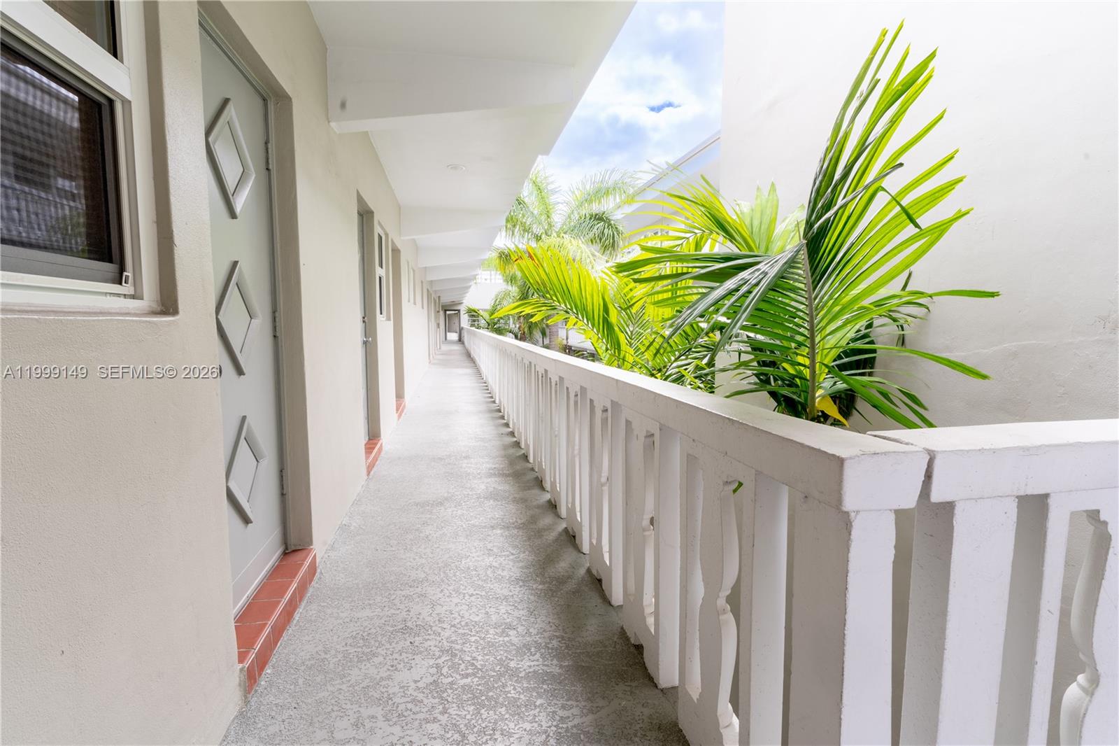 An outdoor apartment building walkway features a white railing, textured concrete flooring, and palm trees — 330 SE 2nd St # 405F, Hallandale Beach FL 33009, HALLANDALE BEACH, FL