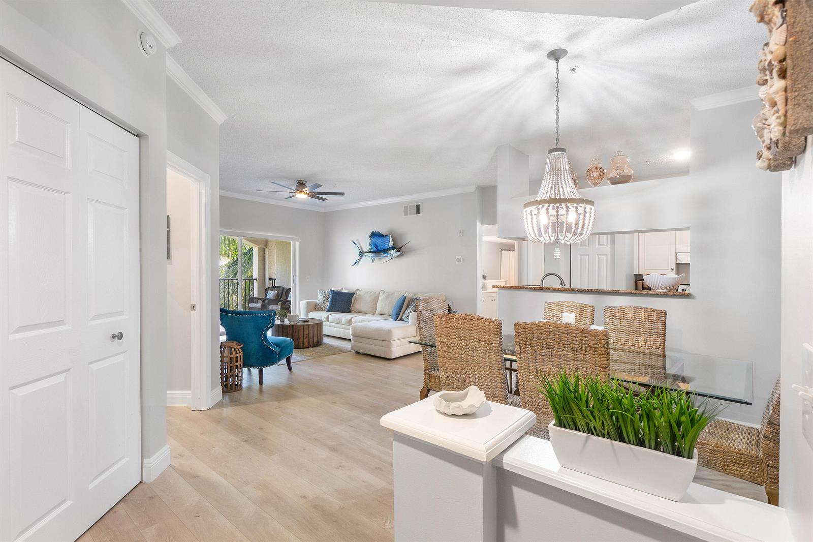 An open-concept living and dining area features light wood floors, wicker chairs, and a chandelier — 2408 Tuscany Way # 2408, Boynton Beach FL 33435, BOYNTON BEACH, FL