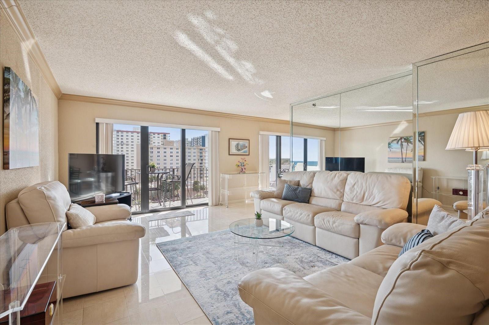 A living room with beige leather sofas, a glass coffee table, and mirrored wall panels — 1201 S Ocean # 704s, Hollywood FL 33019, HOLLYWOOD, FL