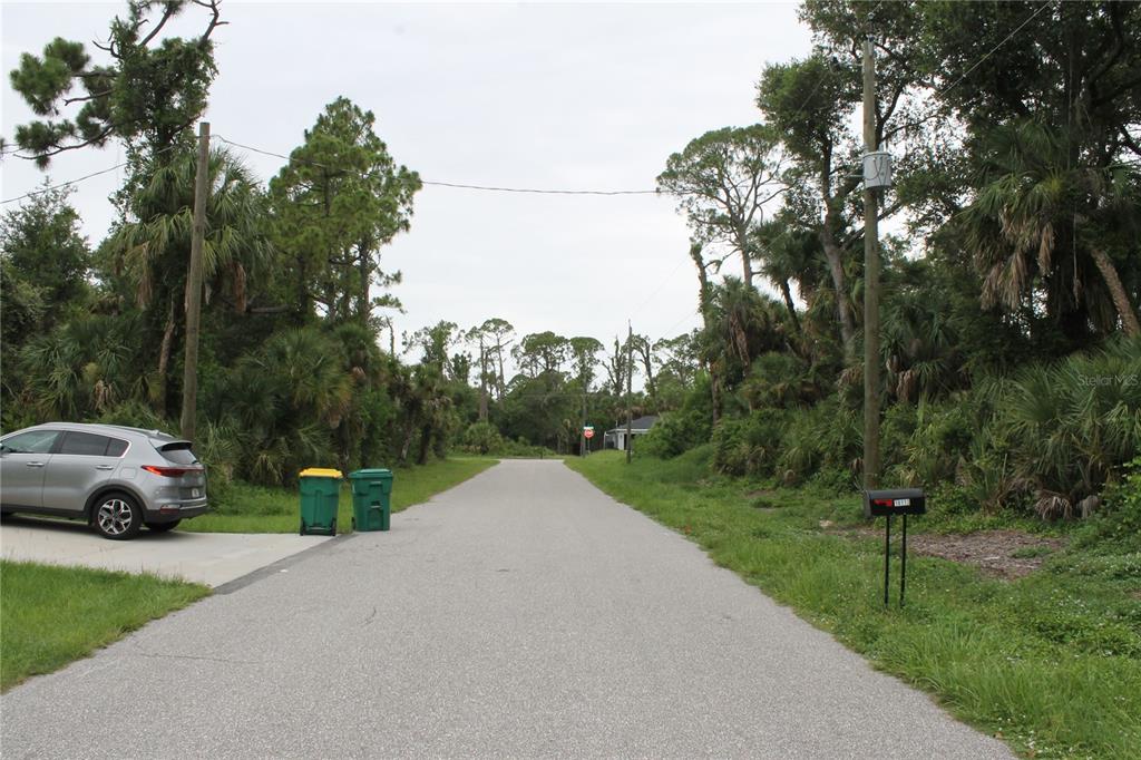 A paved residential street is lined with dense trees and vegetation under a cloudy sky — 18106 SINATRA AVENUE, PORT CHARLOTTE, FL