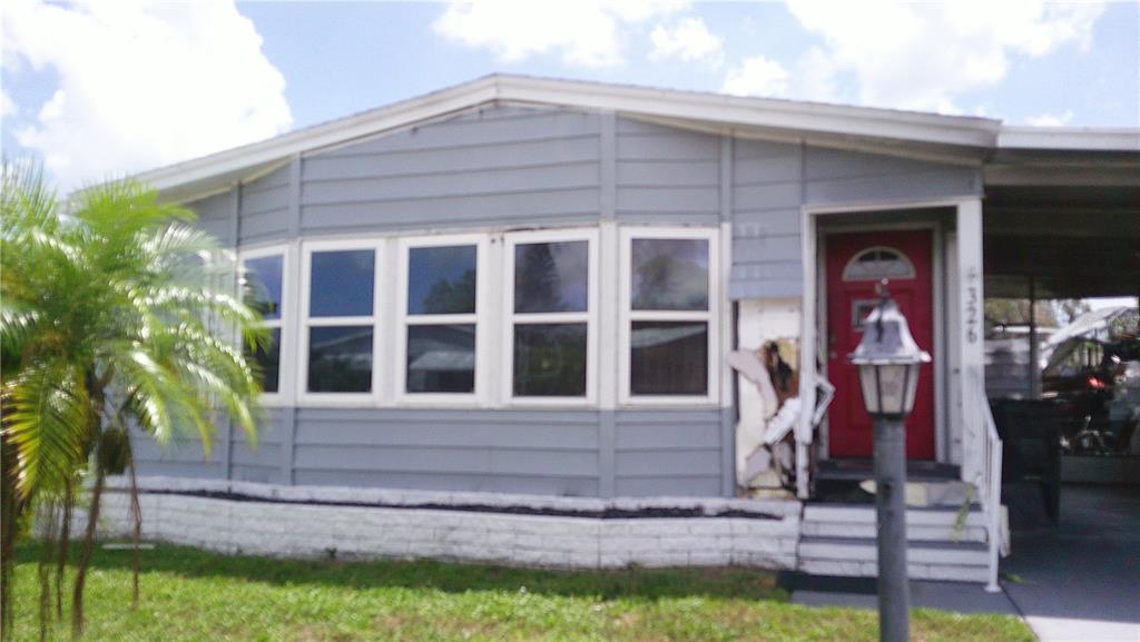 A gray mobile home exterior features a red front door and damaged siding near the entrance — 6710 36TH AVENUE E UNIT 326, PALMETTO, FL
