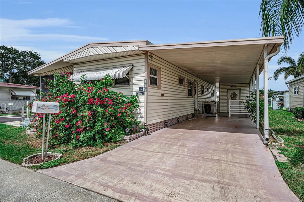A single-story mobile home with beige siding, a carport, and a large flowering bush — 10539 TAHITIAN LANE UNIT 112, LARGO, FL