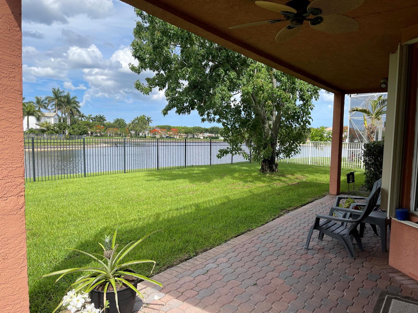 A covered patio with brick pavers overlooks a grassy backyard, a lake, and palm trees — 9828 Stover Way, Wellington FL 33414, WELLINGTON, FL
