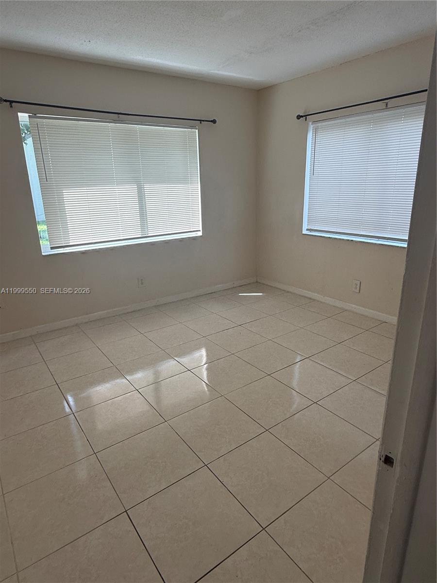 An empty bedroom features light-colored tile flooring, beige walls, and two windows with blinds — 201 SE 6th Ave # 7, Pompano Beach FL 33060, POMPANO BEACH, FL